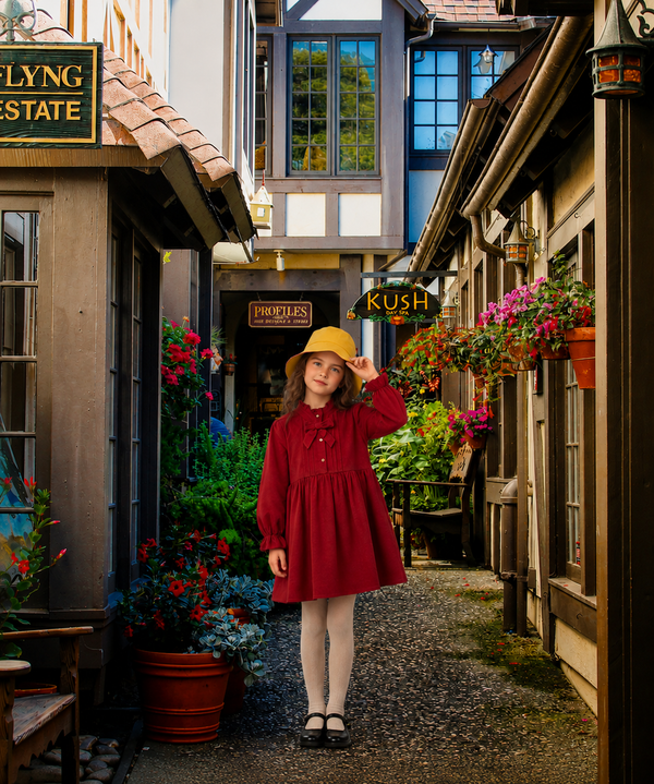 Girl in red corduroy bow dress and yellow hat poses in a quaint village alley, cute toddler girl dresses