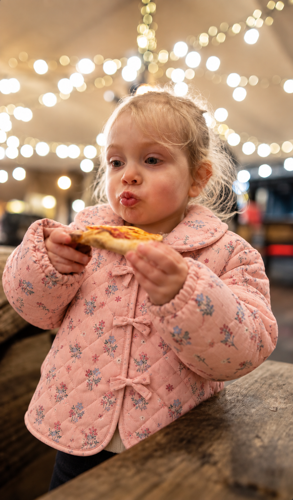 Toddler eating pizza in dusty pink floral jacket under string lights, cozy kids fall clothing