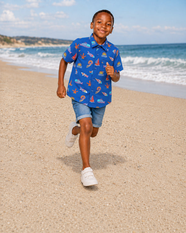 Young boy running on a sandy beach in a blue ocean-print shirt and denim shorts from Kissa beachwear for infants collection