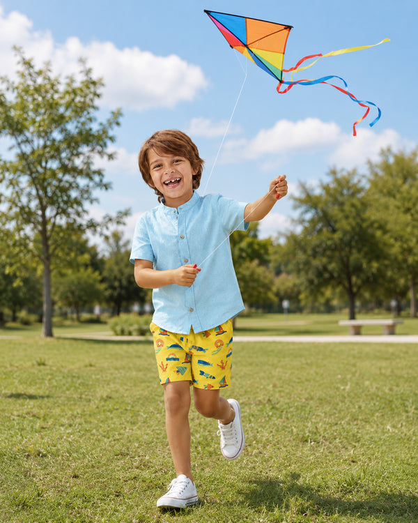Smiling boy wearing yellow ocean-print shorts, part of Kissa beachwear for infants from the Ocean Collection, while flying a colorful kite