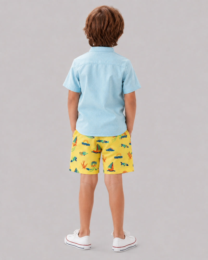 Boy wearing yellow ocean themed shorts with light blue shirt, back view